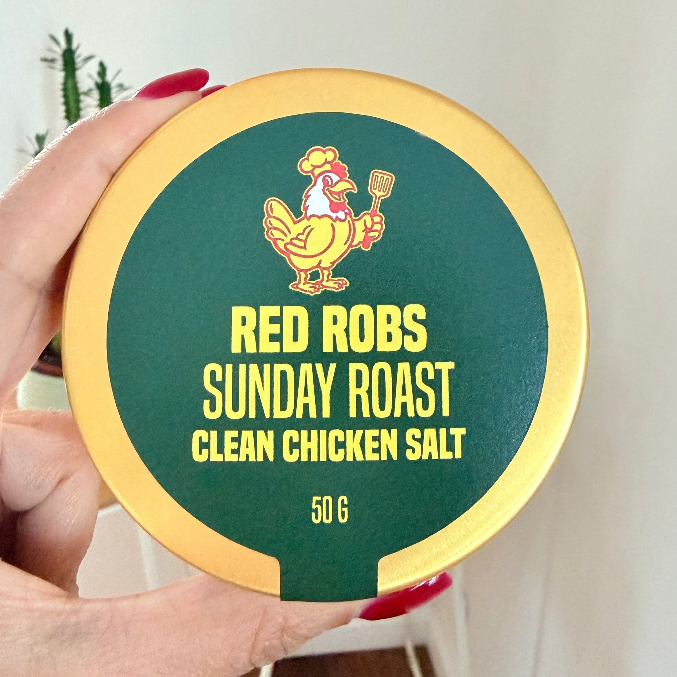A hand with red painted nails holds a round tin of Red Robs Sunday Roast Clean Chicken Salt made with real chicken. The tin has a gold-colored rim and a dark green center with yellow text and a Red Robs Original chicken logo.