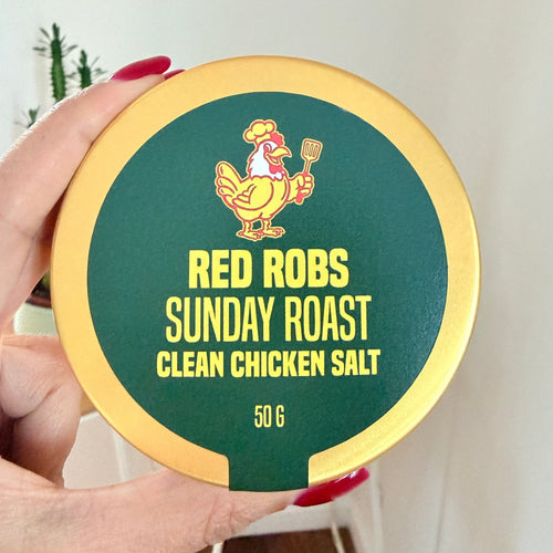 A hand with red painted nails holds a round tin of Red Robs Sunday Roast Clean Chicken Salt made with real chicken. The tin has a gold-colored rim and a dark green center with yellow text and a Red Robs Original chicken logo.